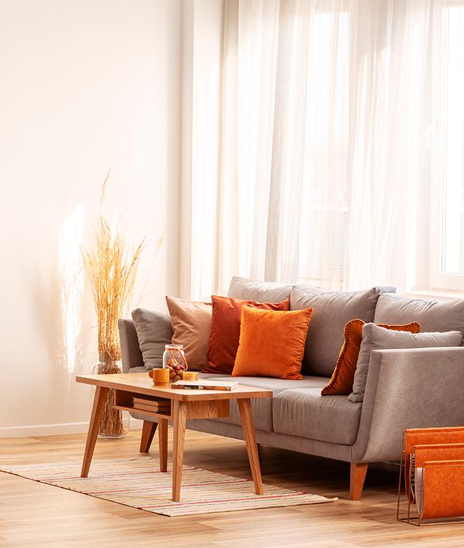 Kodikas olohuone, jossa verhot ikkunan edessä, harmaa sohva jolla eri värisiä sohvatyynyjä, puinen sohvapöytä ja koriste-esineitä.